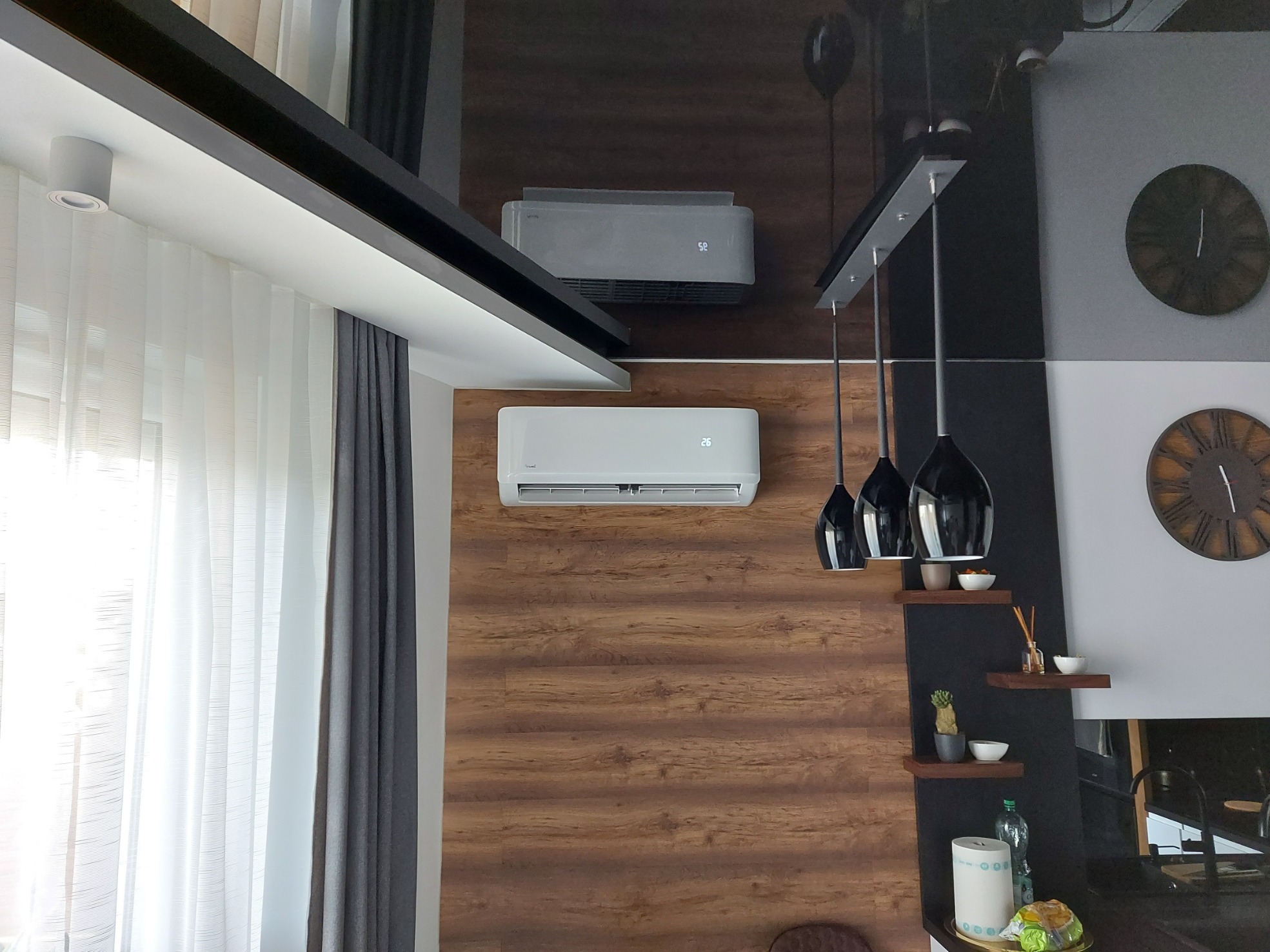 Dwa klimatyzatory zamontowane na ścianie z drewnianą okładziną w nowoczesnym salonie z czarnymi lampami wiszącymi i dekoracjami na półkach.