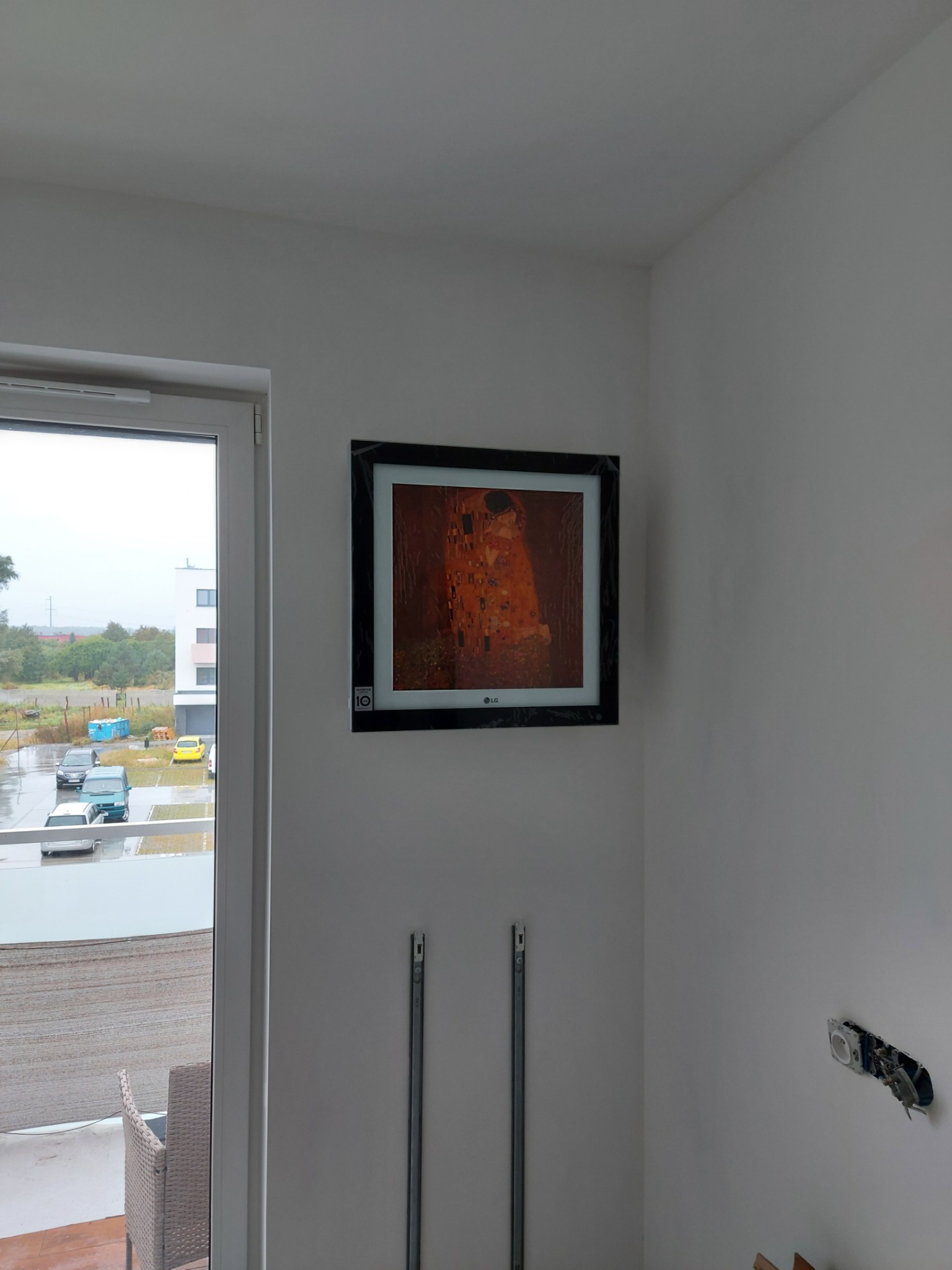 Wnętrze pomieszczenia z widokiem na zewnątrz przez okno balkonowe, na ścianie wisi telewizor z obrazem Gustava Klimta 'Pocałunek', widoczne metalowe szyny montażowe na ścianie i niezabezpieczone...