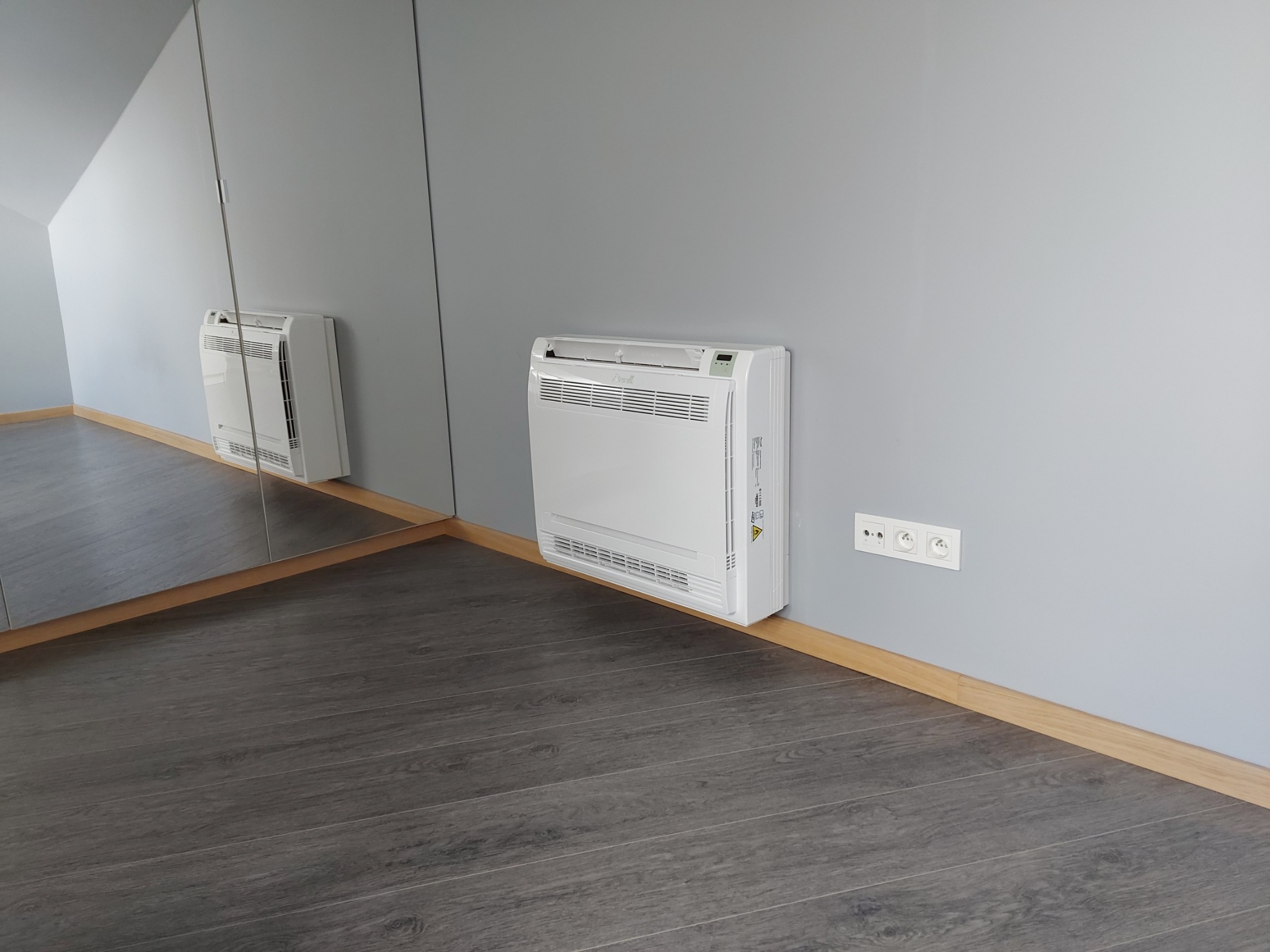 Biały klimatyzator ścienny zamontowany w rogu pokoju z szarą ścianą i ciemną podłogą, widoczny również w lustrzanym odbiciu.