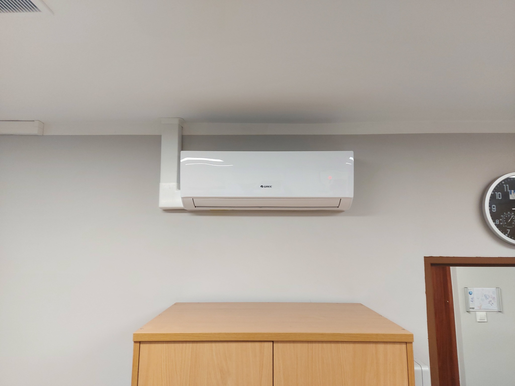 Biały klimatyzator marki Gree zamontowany na ścianie nad drewnianą komodą w biurze, widoczny zegar i odbicie w lustrze.