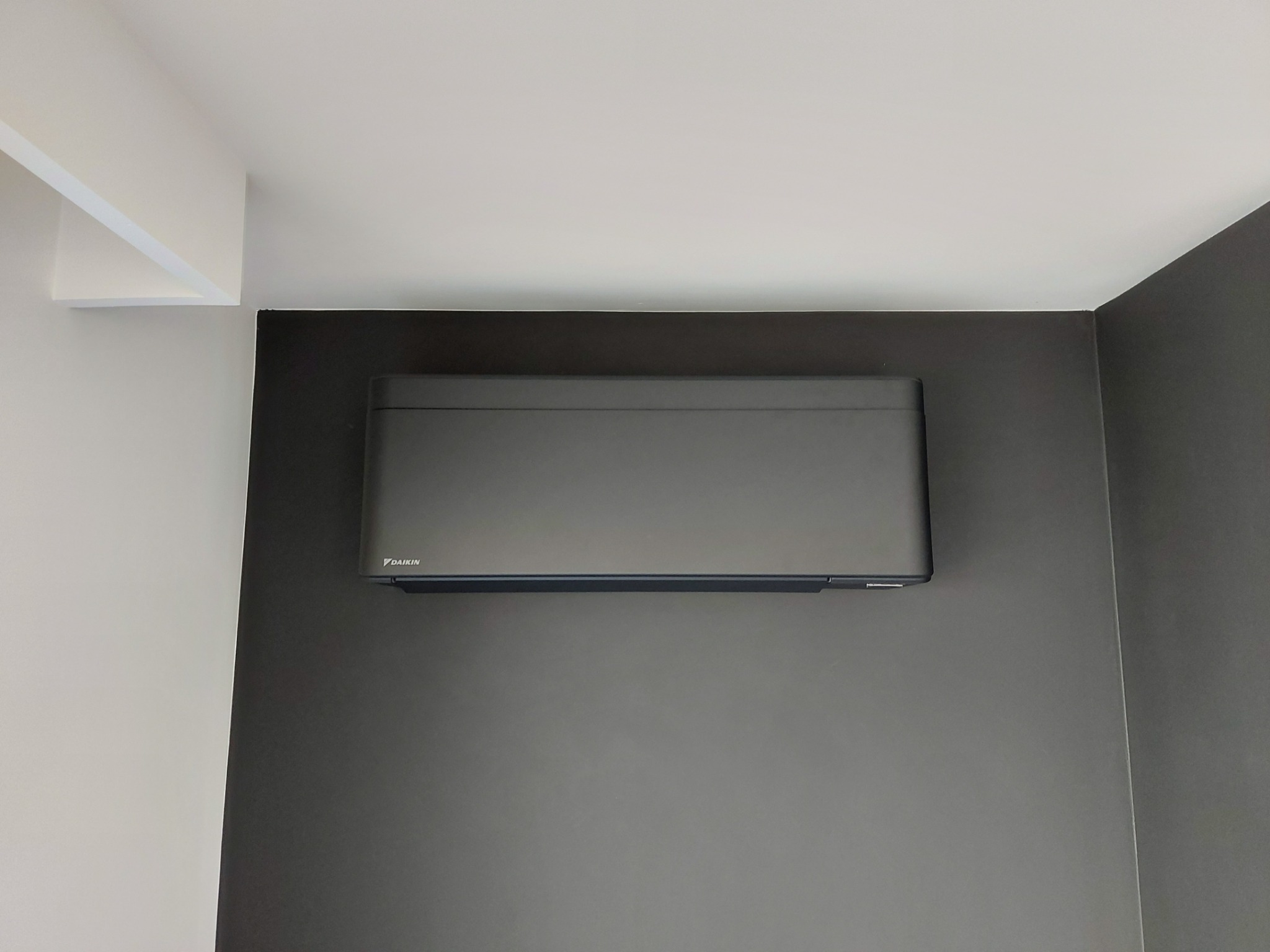 Czarny klimatyzator Daikin zamontowany na ciemnoszarej ścianie w rogu pomieszczenia z białym sufitem i fragmentem białej ściany.