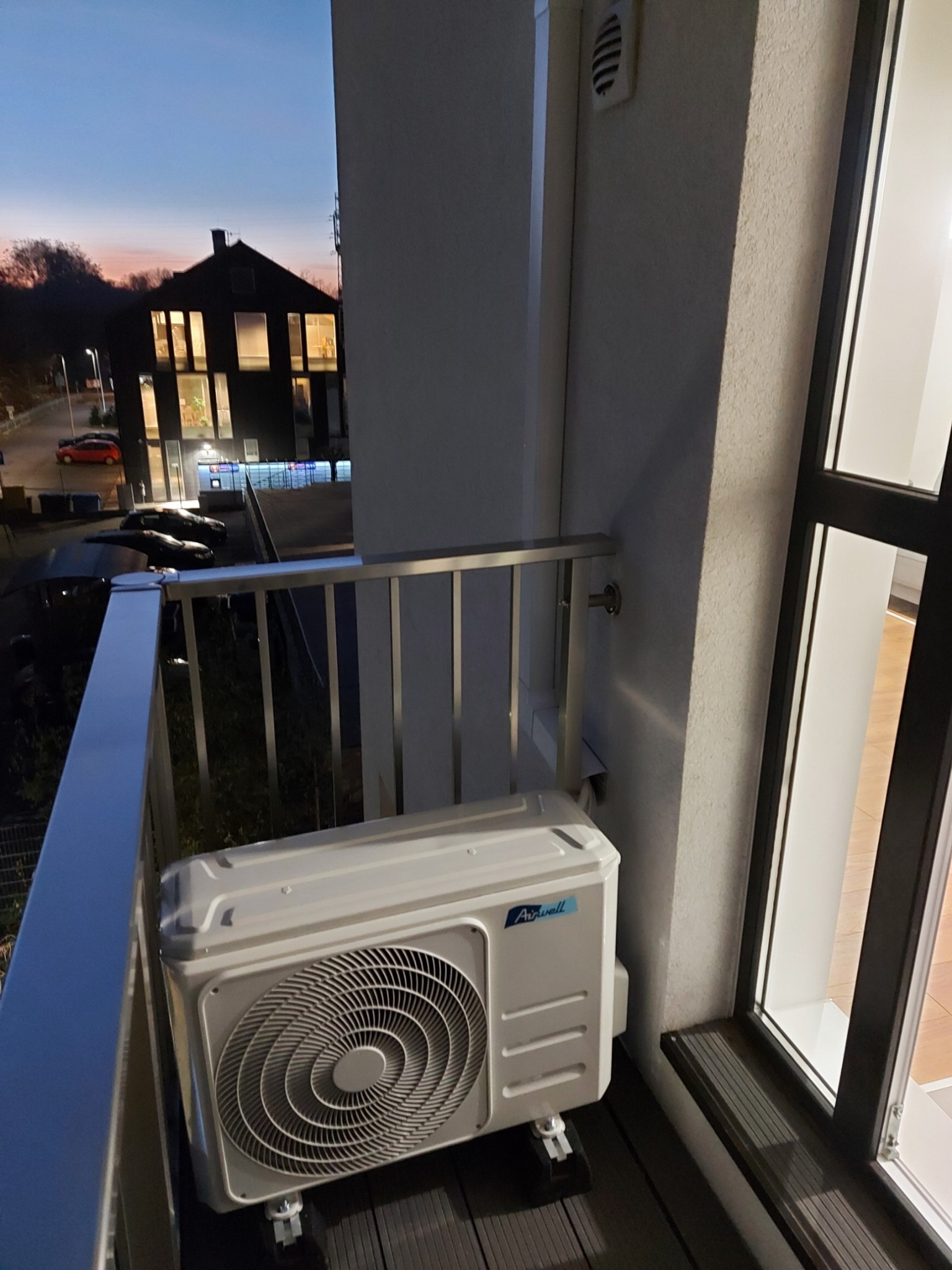 Jednostka zewnętrzna klimatyzatora marki Airwell zamontowana na balkonie, widok na budynek mieszkalny o zmierzchu.