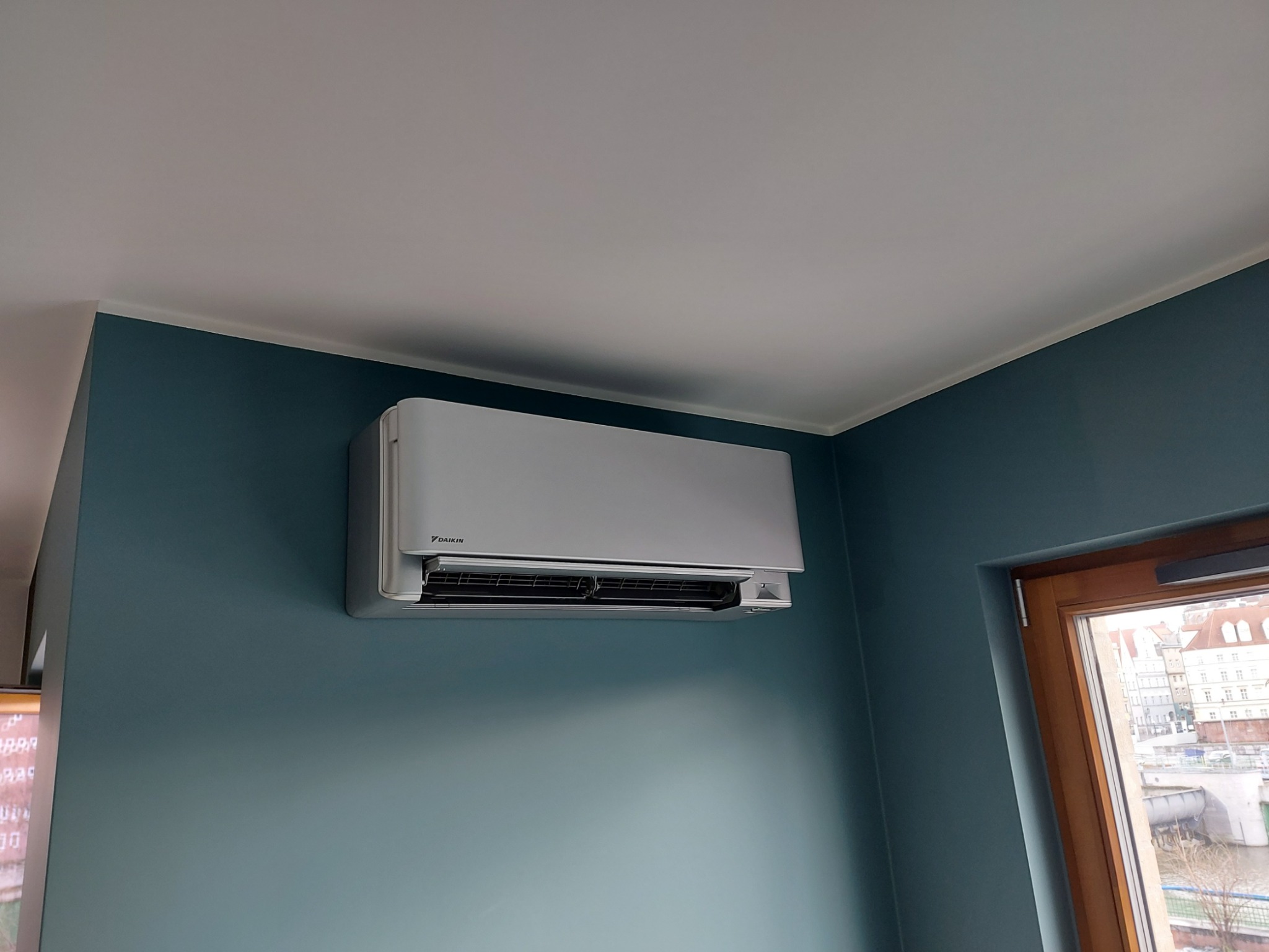 Szary klimatyzator Daikin zamontowany na błękitnej ścianie w pomieszczeniu z oknem, widok na miasto i rzekę.