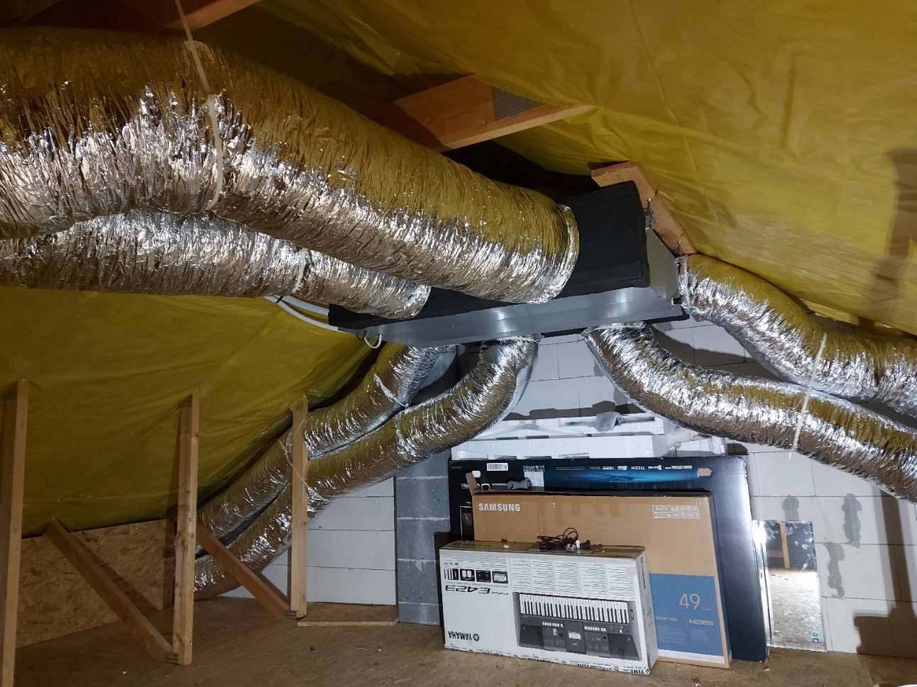 Instalacja wentylacji w przestrzeni poddasza: srebrne, elastyczne rury wentylacyjne rozchodzą się od centralnej jednostki, widoczne elementy konstrukcyjne dachu i kartony z elektroniką w tle.