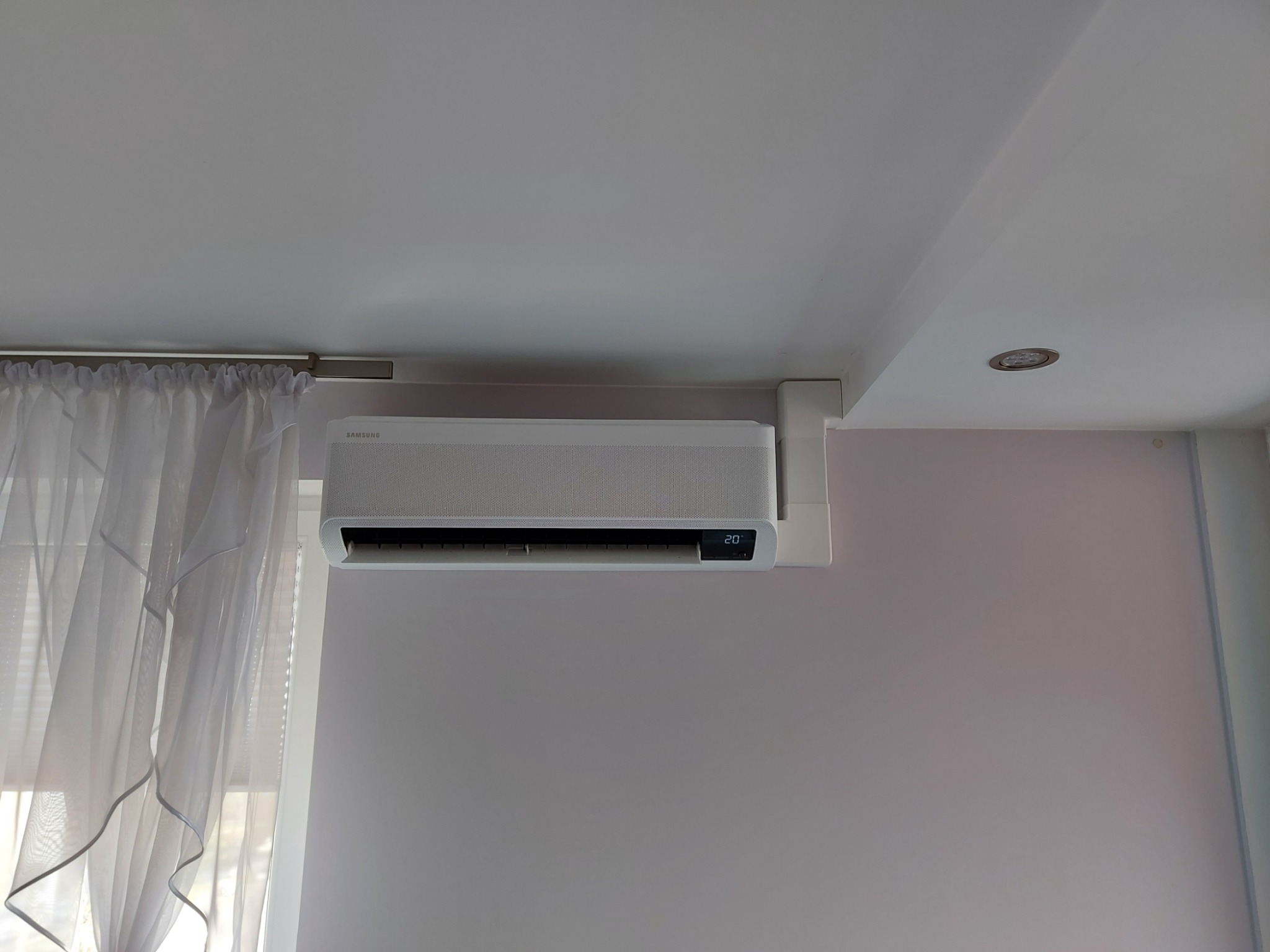 Biała jednostka klimatyzacji Samsung zamontowana na ścianie w pokoju z jasnymi zasłonami, wyświetlająca temperaturę 20 stopni Celsjusza.