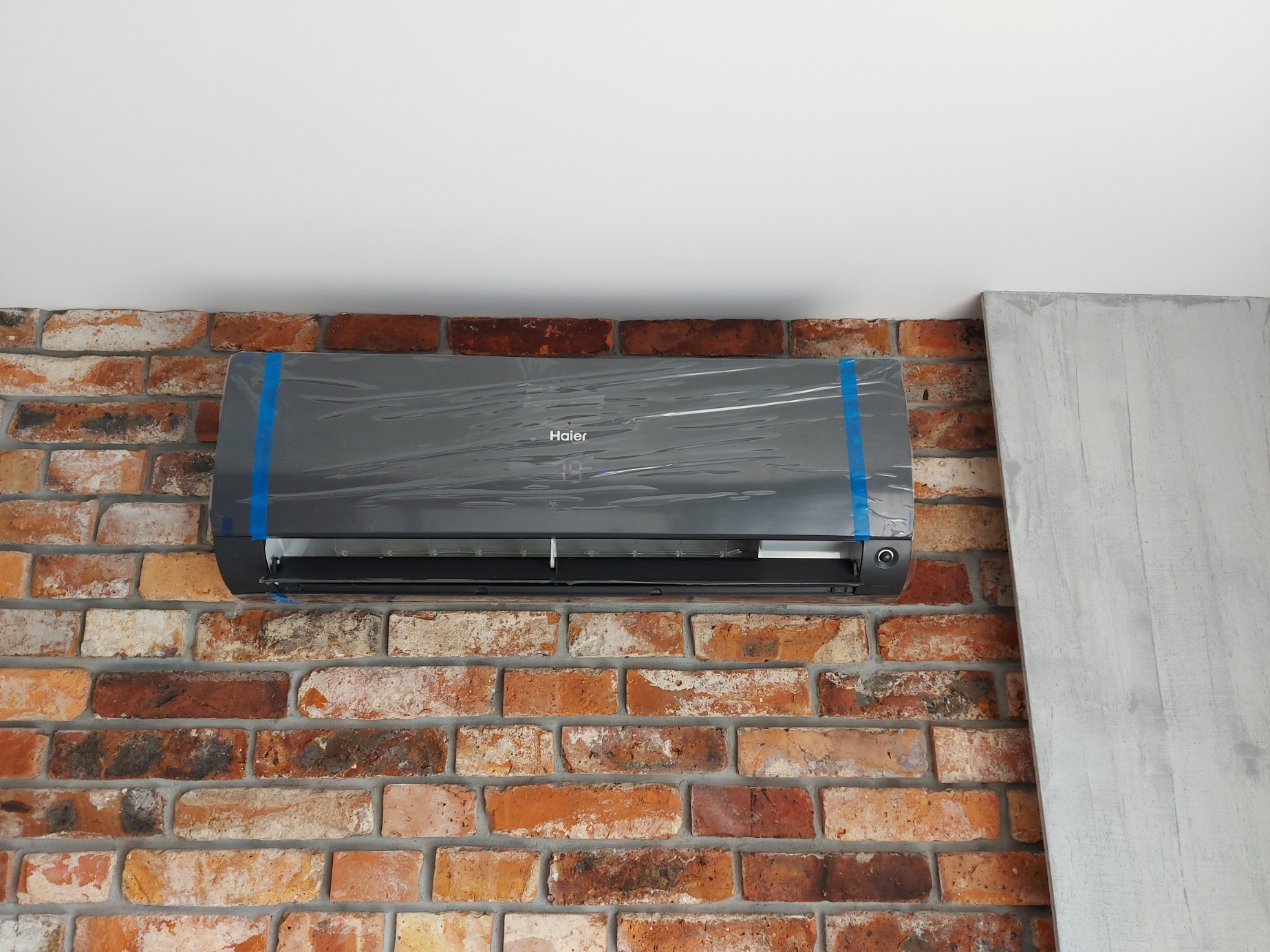 Klimatyzator Haier zamontowany na ceglanej ścianie, jeszcze w folii ochronnej z niebieską taśmą, widoczny fragment betonowej płyty na ścianie.