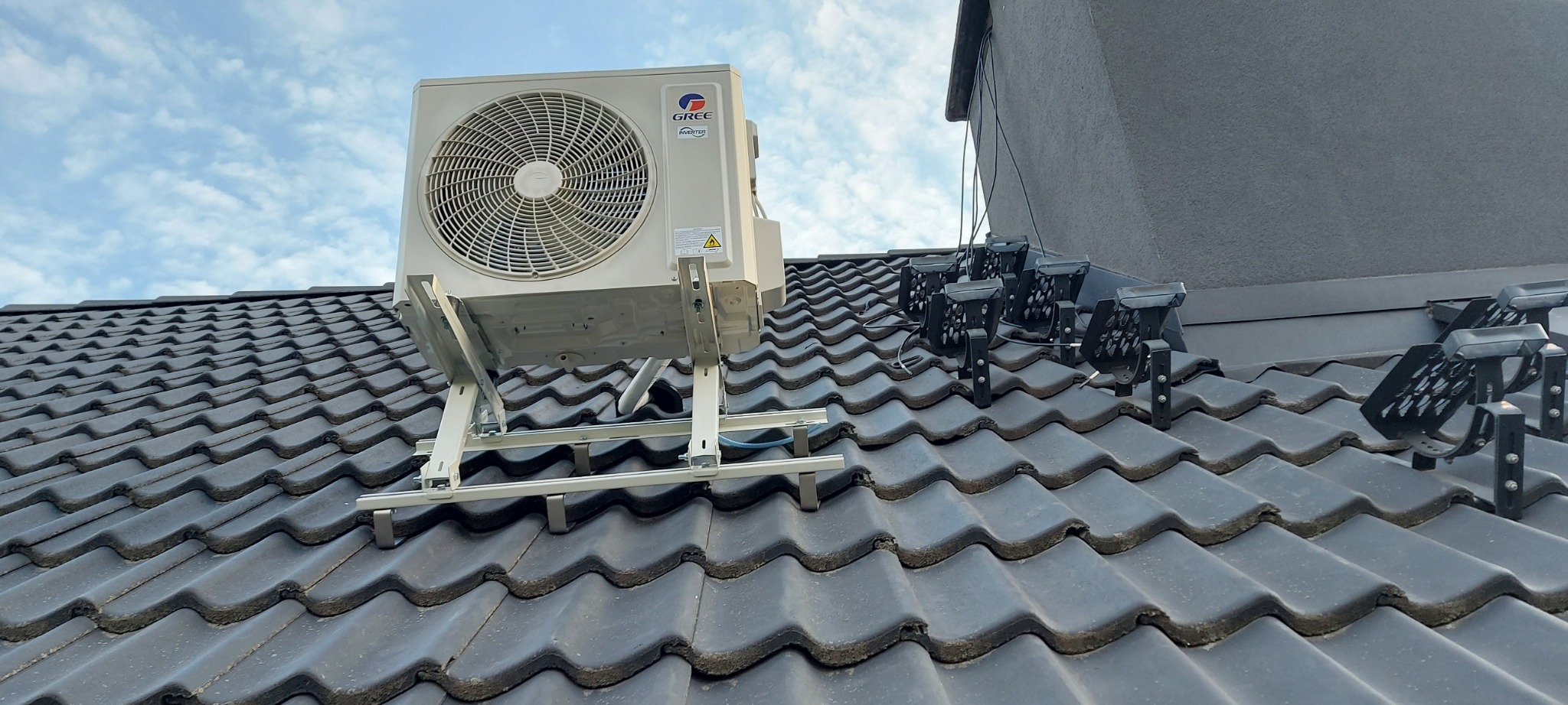 Biała jednostka zewnętrzna klimatyzatora marki Gree zamontowana na metalowym stelażu na ciemnoszarym dachu z dachówki ceramicznej, z widocznymi elementami instalacji fotowoltaicznej w tle.