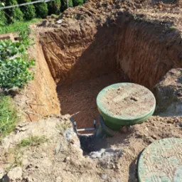 Wykop pod szambo z widocznymi betonowymi kręgami oraz metalową drabinką ułatwiającą zejście, świeżo usypana ziemia wokół wykopu.