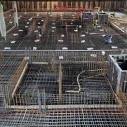 Siatka zbrojeniowa fundamentu z dystansami, szalunkami i pracownikami w kaskach w tle, widok z góry.