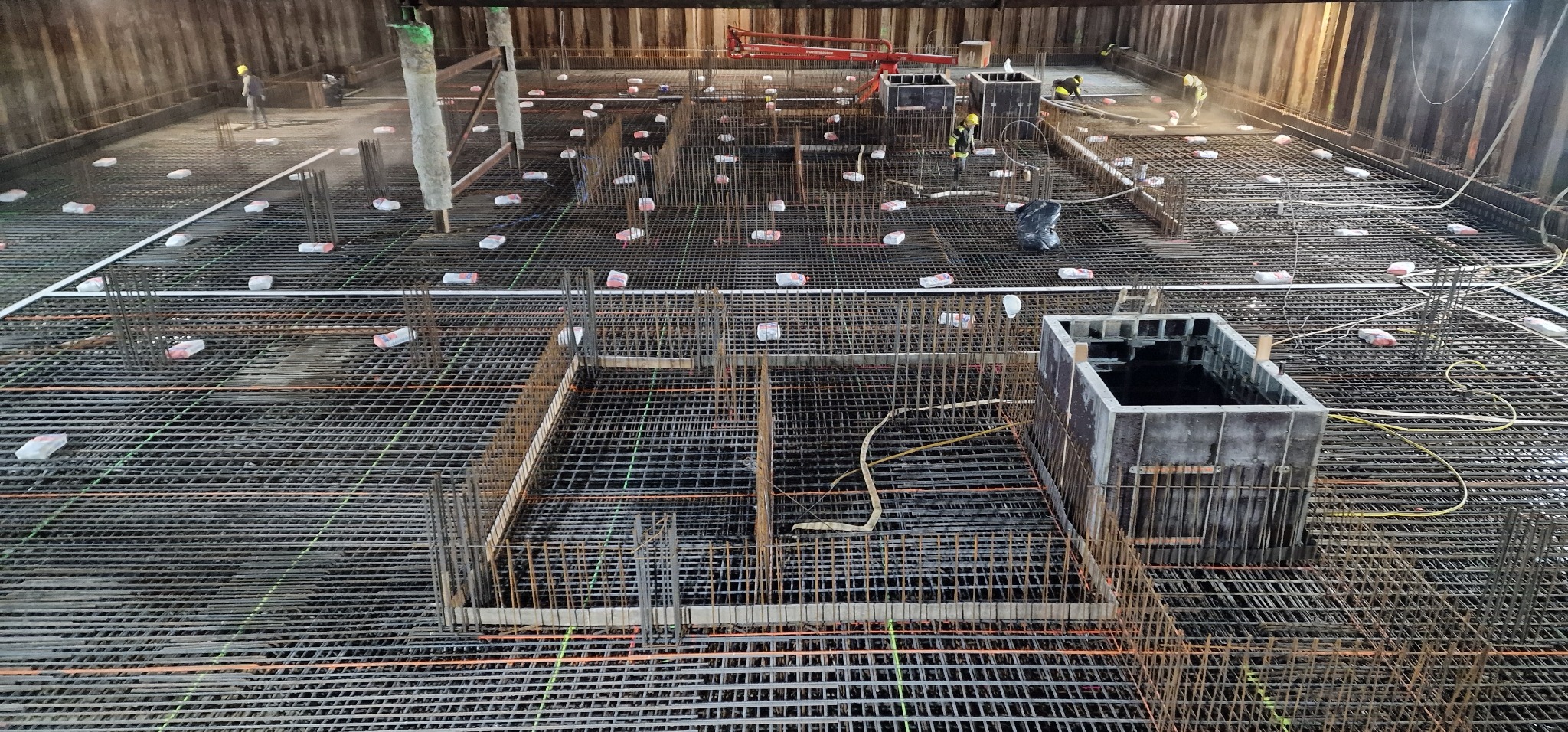 Siatka zbrojeniowa fundamentu z dystansami, szalunkami i pracownikami w kaskach w tle, widok z góry.
