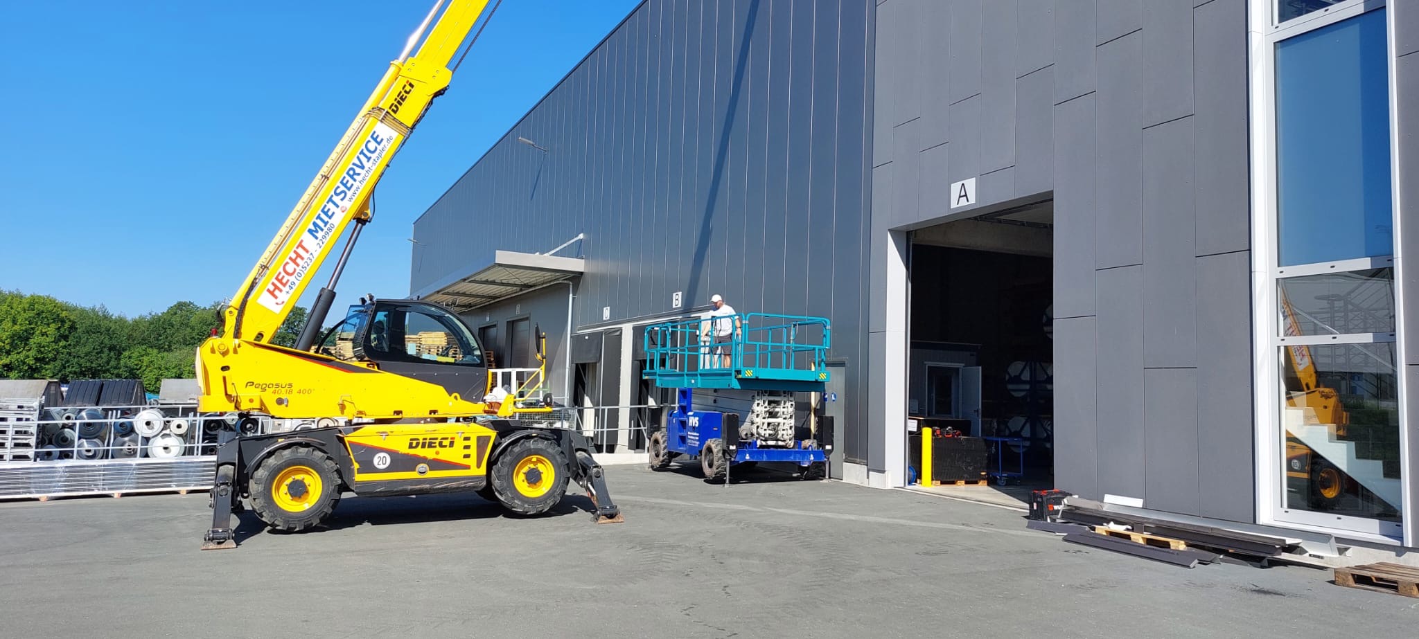 Żółty podnośnik teleskopowy Dieci i niebieska platforma podnośnikowa przy szarej hali z płyt warstwowych, pracownik na platformie przy wejściu oznaczonym literą A.