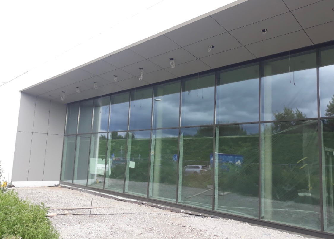 Nowoczesny budynek z przeszkloną fasadą, widoczne elementy konstrukcyjne dachu i instalacji elektrycznej, teren wokół budynku w trakcie prac wykończeniowych.