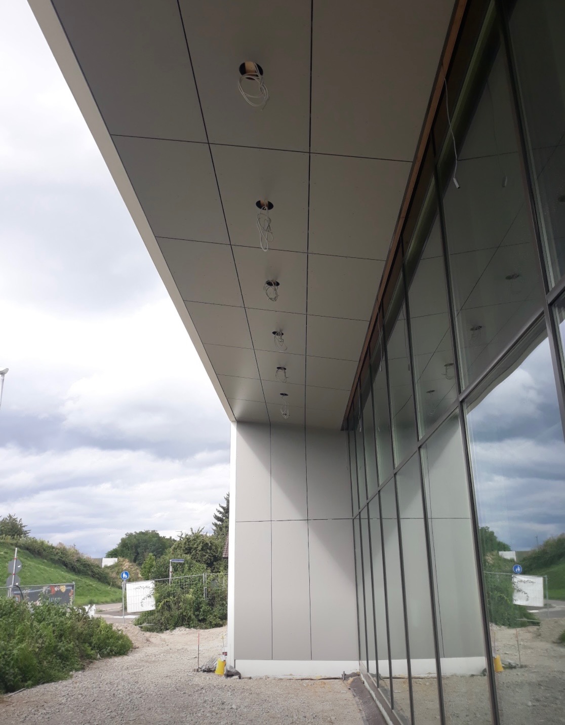 Montaż płyt warstwowych na elewacji budynku z widocznymi otworami na oświetlenie i dużymi oknami, widok z zewnątrz na tle pochmurnego nieba.