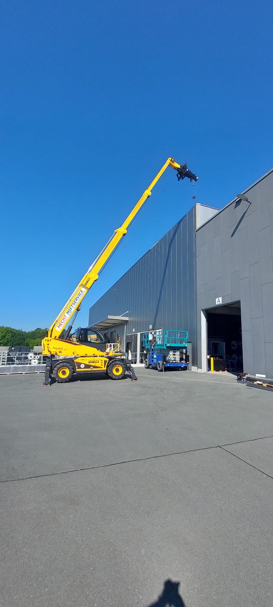 Żółty podnośnik teleskopowy Dieci z wysięgnikiem unosi element konstrukcyjny na tle szarej hali w miejscowości Biała, obok widoczny podnośnik nożycowy.