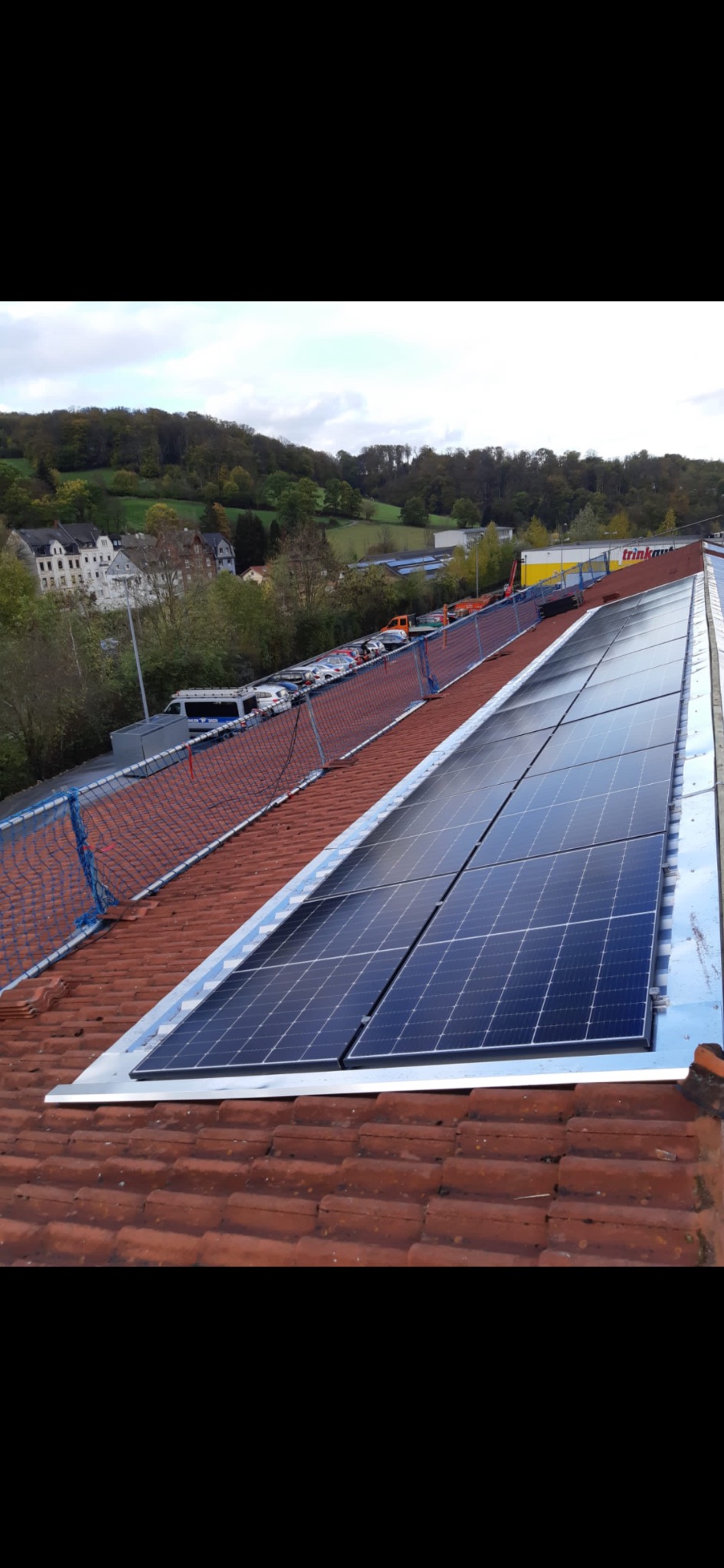 Panele słoneczne zamontowane na dachu z czerwonej dachówki, widoczna siatka zabezpieczająca, w tle budynki i zieleń.