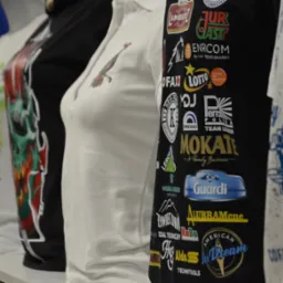 Rząd manekinów z różnymi wzorami na koszulkach, od kolorowych grafik po koszulki polo z haftem i czarne koszulki z nadrukami logo różnych firm.