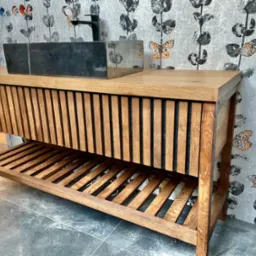 Drewniana szafka łazienkowa z blatem i umywalką z czarnego kamienia, widoczne ryflowane fronty i półka dolna, na tle ściany w tapetę z motywem motyli.