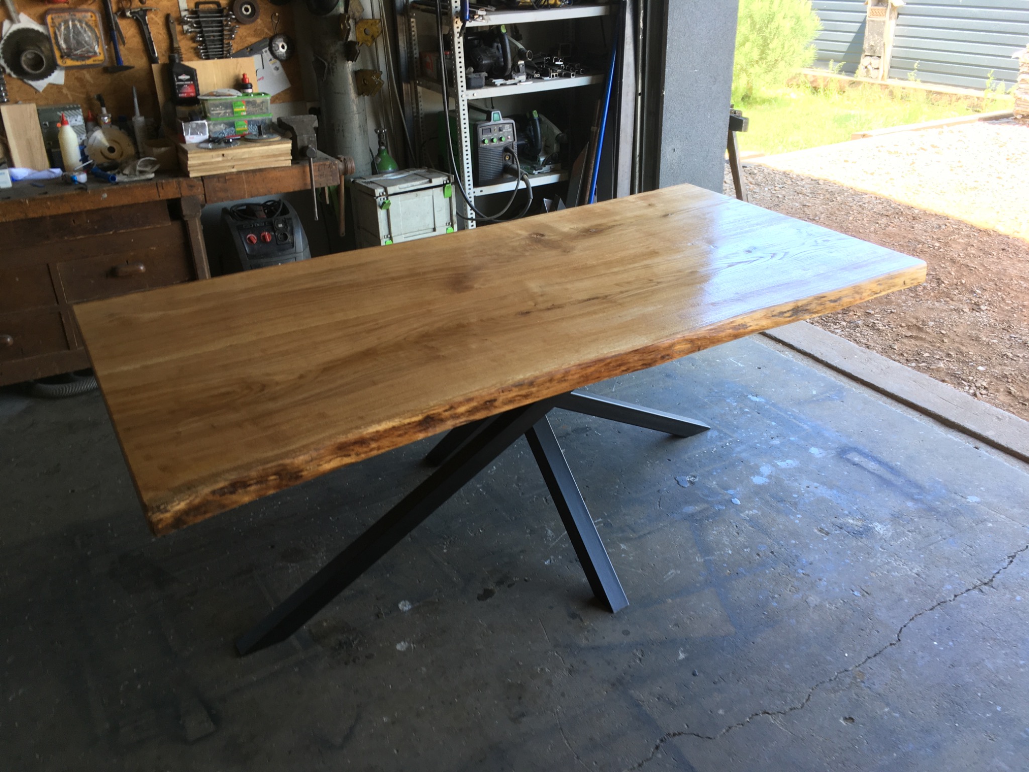 Blat stołu z naturalną krawędzią drewna, osadzony na metalowych nogach w kształcie litery X, widoczny w warsztacie z narzędziami i spawarką w tle.