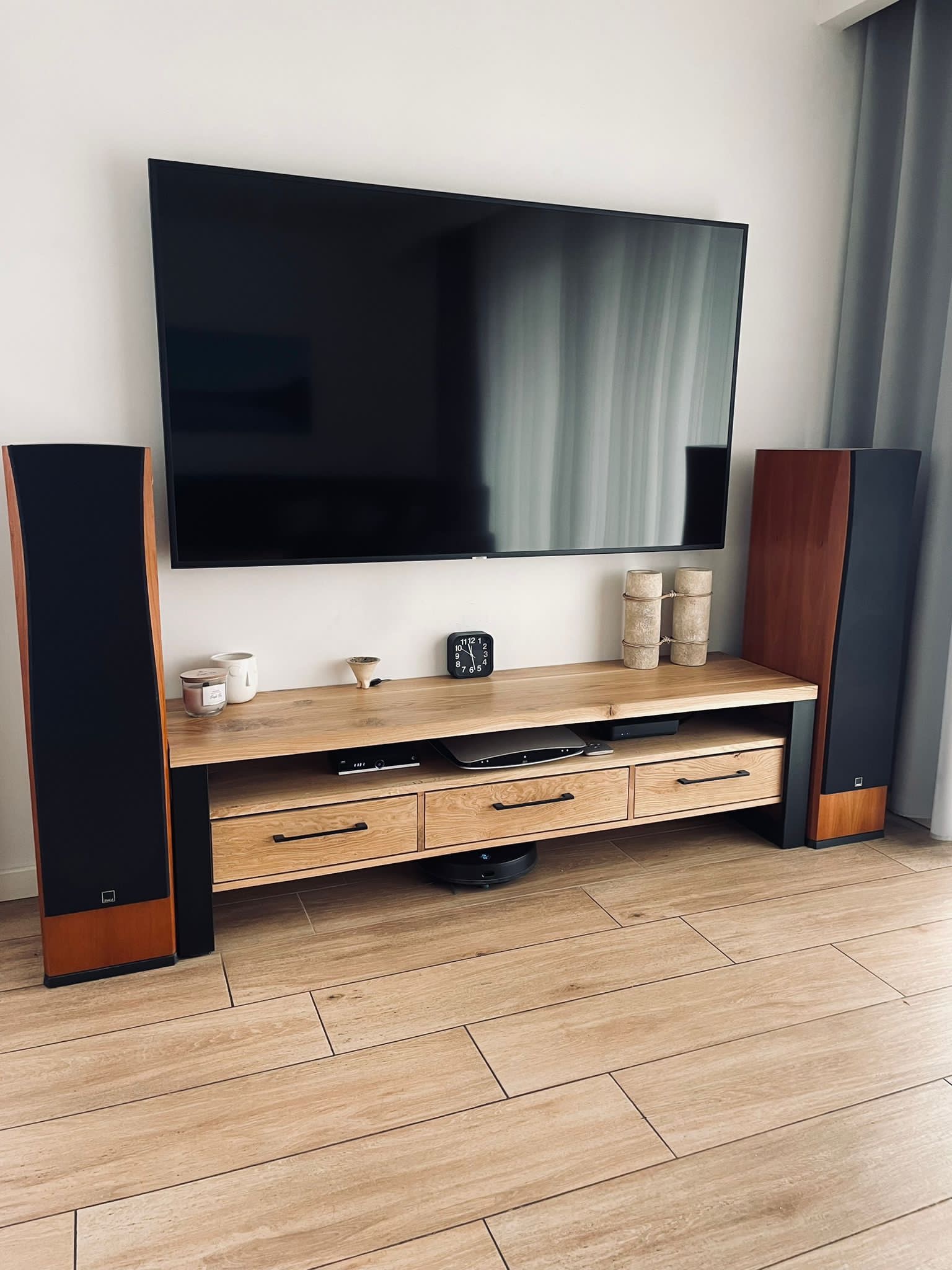 Drewniana szafka RTV z trzema szufladami i otwartymi półkami, umieszczona między dwoma kolumnami głośnikowymi, pod wiszącym telewizorem na białej ścianie. Na blacie szafki dekoracje i zegar, a...