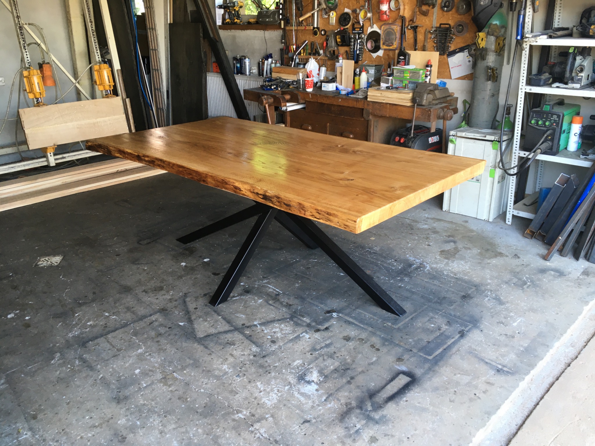 Jasny, lakierowany blat drewniany z naturalną krawędzią na metalowych nogach w kształcie litery X, widoczny w warsztacie stolarskim z narzędziami i materiałami w tle.