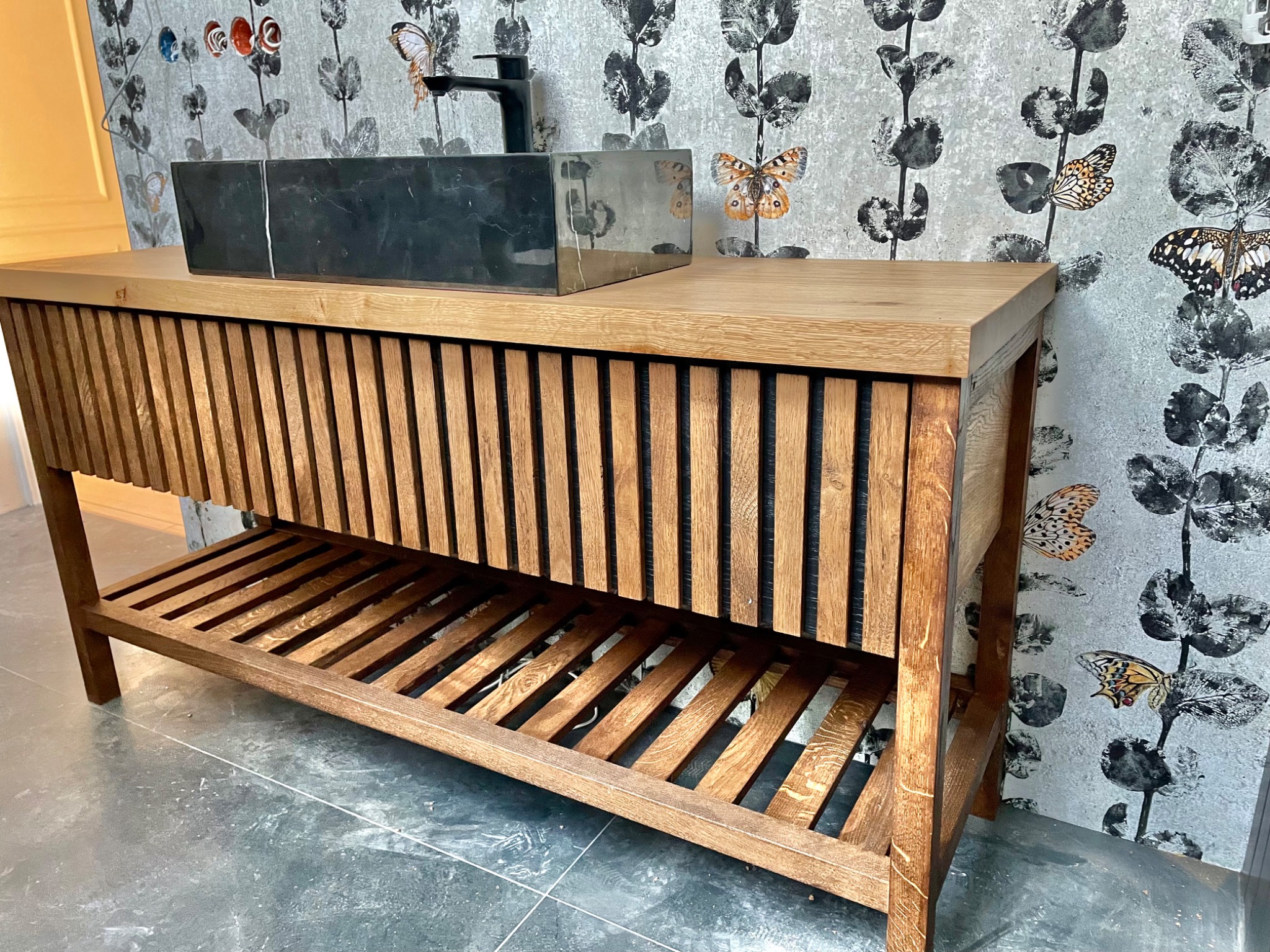Drewniana szafka łazienkowa z blatem i umywalką z czarnego kamienia, widoczne ryflowane fronty i półka dolna, na tle ściany w tapetę z motywem motyli.