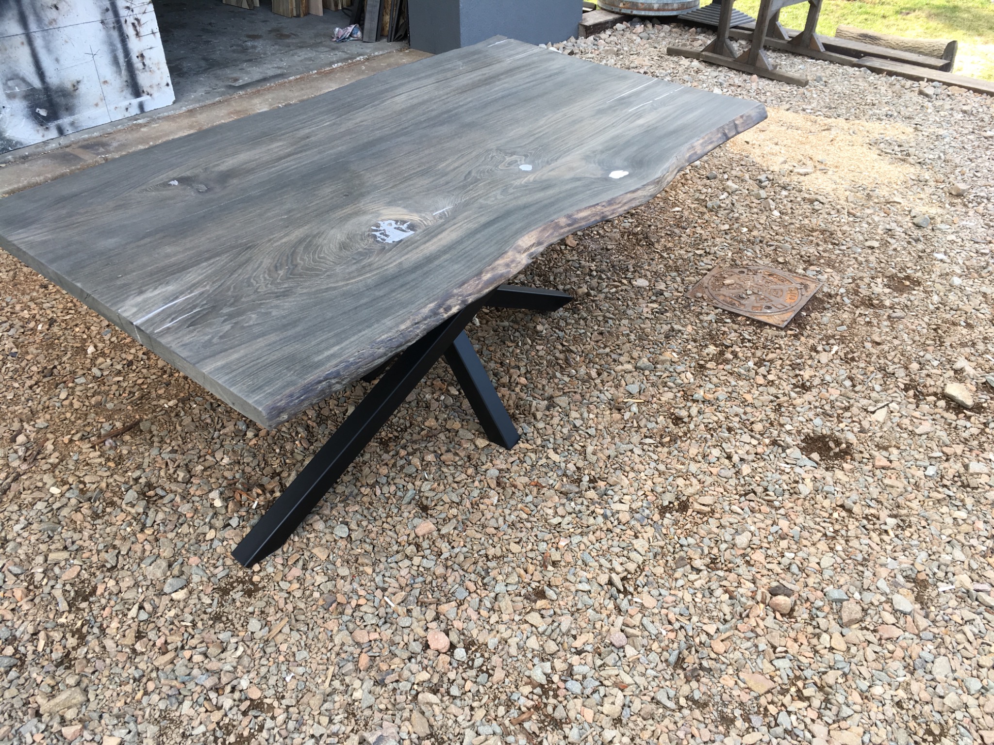 Stół z blatem z surowego drewna w odcieniach szarości, z widocznymi naturalnymi krawędziami i wypełnionymi żywicą ubytkami, osadzony na czarnej, metalowej podstawie w kształcie litery X, ustawiony...