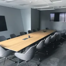 Nowoczesna sala konferencyjna z długim, jasnym stołem i szarymi krzesłami na kółkach, czarny telewizor na ścianie, minimalistyczne oświetlenie sufitowe.