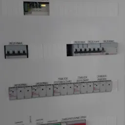 Rozdzielnica elektryczna z opisami obwod&oacute;w: licznik energii PV, rezerwa, tablica fotowoltaiki, kotłowni, parter, zabezpieczenie PPOŻ oraz moduły zabezpieczeń. Widoczne diody sygnalizacyjne.