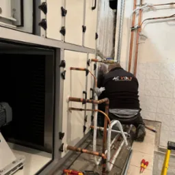 Instalator w czarnej bluzie z logo firmy AC VOLT instaluje miedziane rury do białej centrali wentylacyjnej w pomieszczeniu wyłożonym kafelkami, widoczne narzędzia i drabina.
