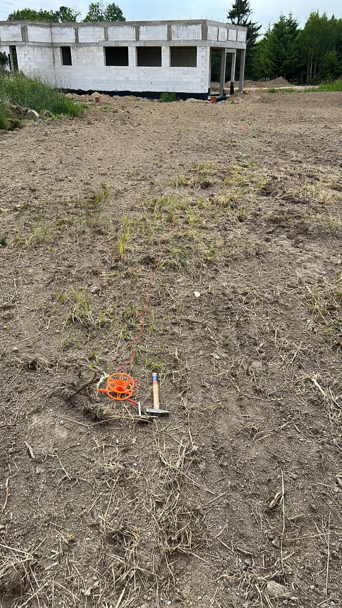 Pomarańczowa szpula z linką, młotek i szpilka wbite w ziemię, wyznaczające trasę instalacji odgromowej na tle surowego budynku.