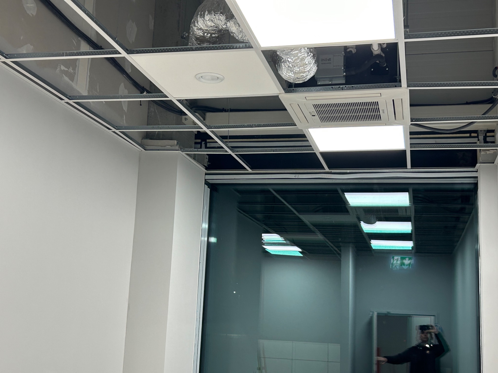 Widok na sufit podwieszany z widocznymi elementami instalacji klimatyzacji, w tym kanałami wentylacyjnymi i jednostką wewnętrzną, oraz oświetleniem LED, częściowo zasłonięty przez konstrukcję...