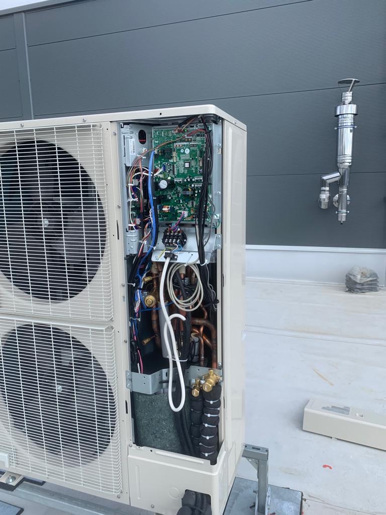 Otwarta obudowa zewnętrznej jednostki klimatyzacji z widoczną elektroniką, miedzianymi rurami i okablowaniem, zamontowana na dachu budynku z szarą elewacją.