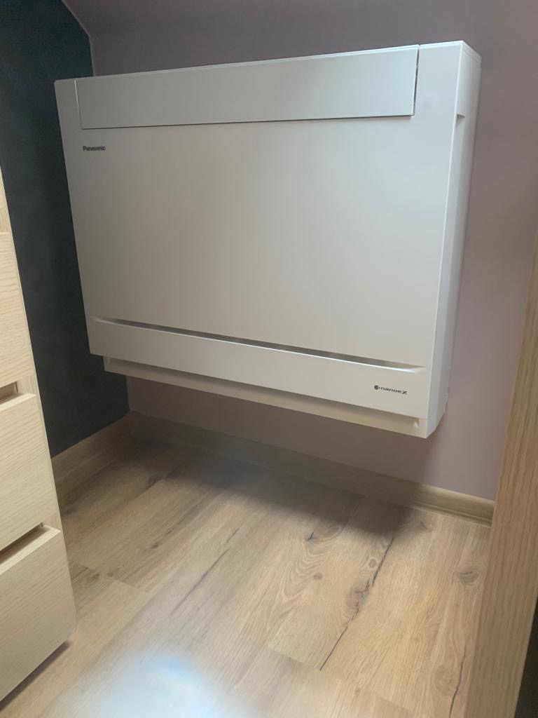 Biała, przypodłogowa jednostka klimatyzacyjna Panasonic zamontowana na ścianie w pomieszczeniu z drewnianą podłogą, widoczna z perspektywy lekko z dołu.
