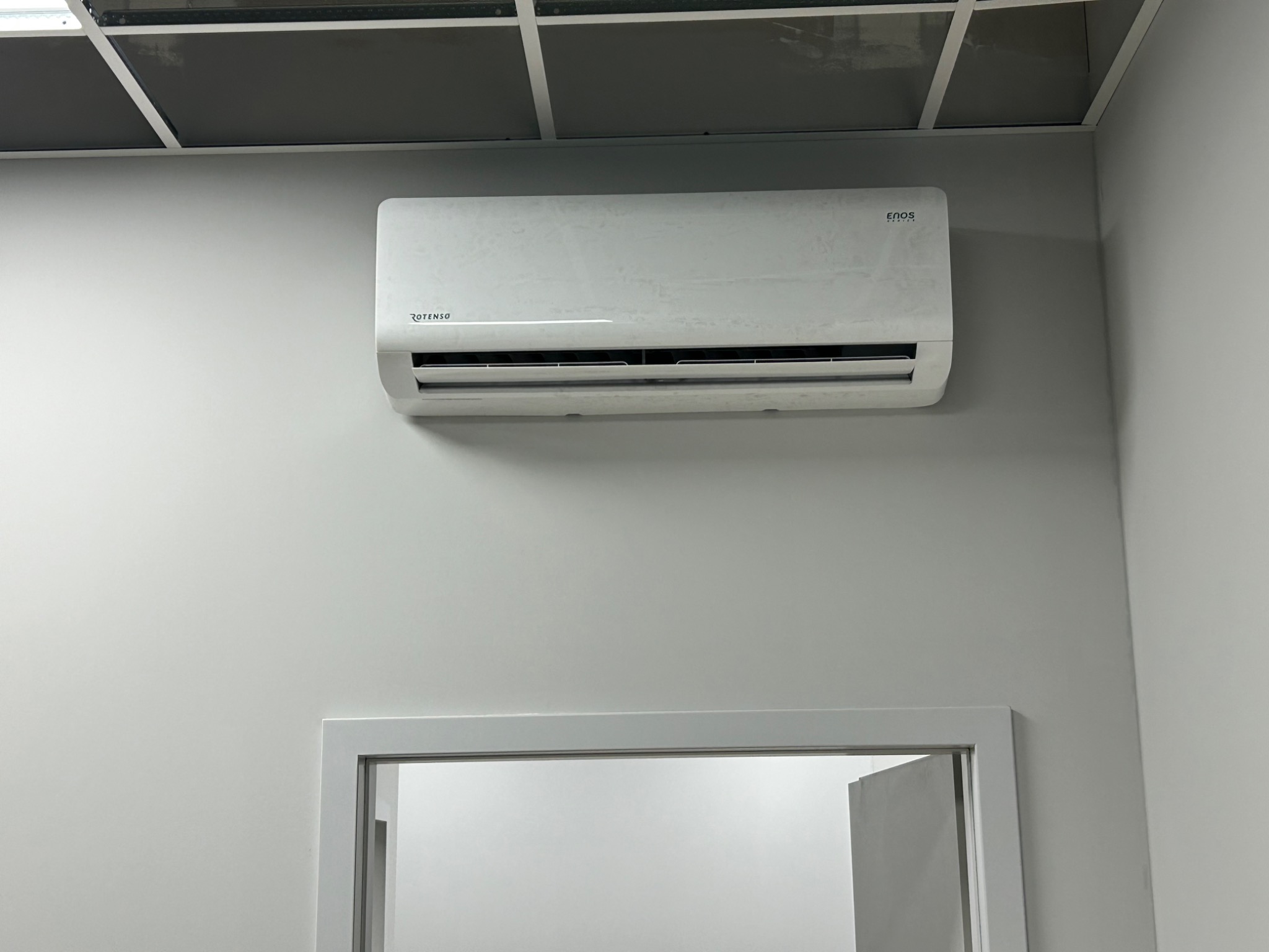 Biała jednostka klimatyzacyjna marki Rotenso zamontowana na szarej ścianie w biurze, widoczny fragment sufitu kasetonowego i ościeżnica drzwi.