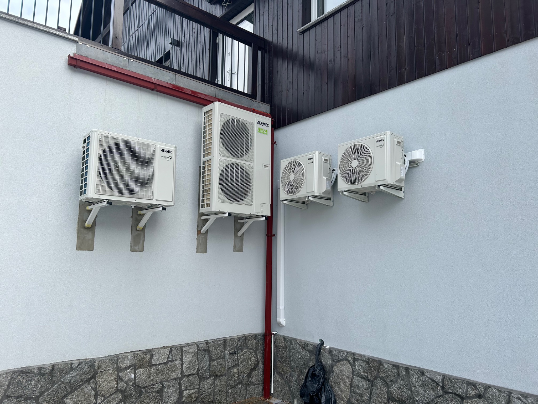 Zewnętrzne jednostki klimatyzacyjne Aermec zamontowane na jasnoszarej elewacji domu z ciemnobrązową drewnianą górną częścią i kamienną podmurówką.