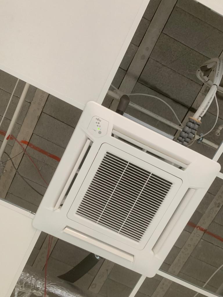 Biała, sufitowa kaseta klimatyzacyjna marki Fujitsu zamontowana w biurze z widoczną instalacją elektryczną i rurami wentylacyjnymi w suficie podwieszanym.