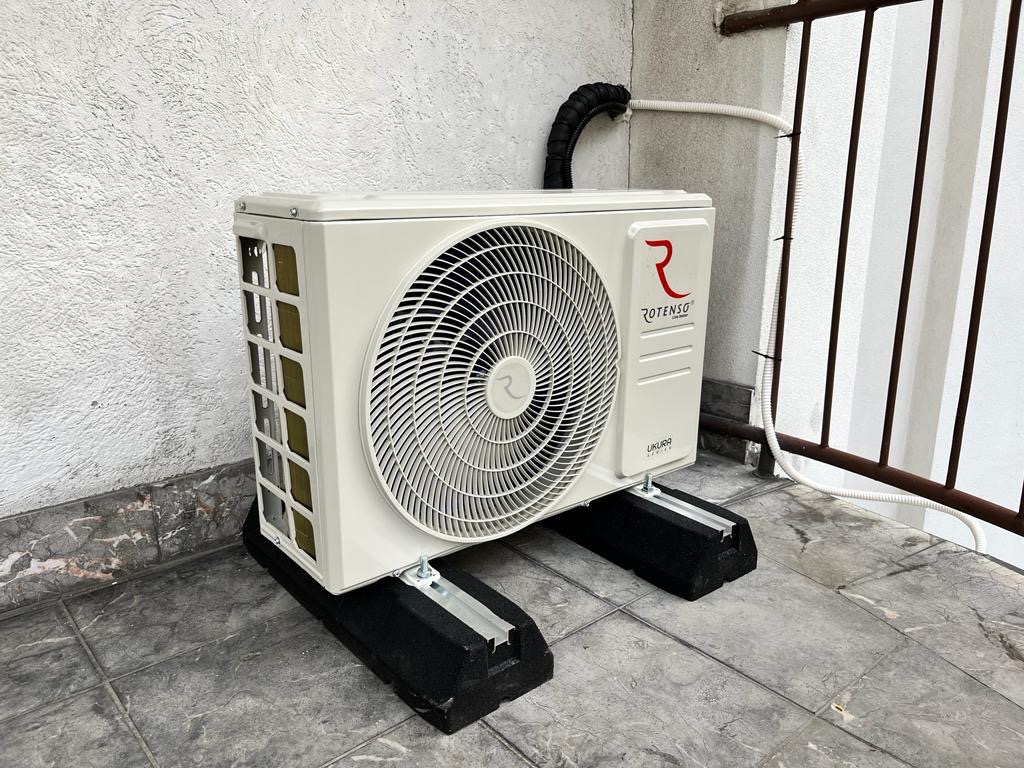 Biała jednostka zewnętrzna klimatyzatora Rotenso ustawiona na czarnych podkładkach antywibracyjnych na balkonie, z widocznymi przewodami i fragmentem balustrady.