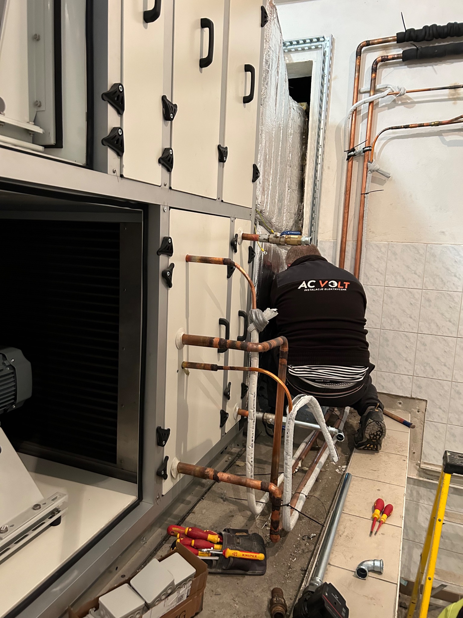 Instalator w czarnej bluzie z logo firmy AC VOLT instaluje miedziane rury do białej centrali wentylacyjnej w pomieszczeniu wyłożonym kafelkami, widoczne narzędzia i drabina.