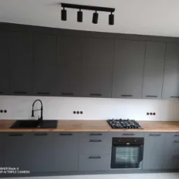 Nowoczesna kuchnia w odcieniach szarości z drewnianym blatem, czarną armaturą i oświetleniem punktowym, widoczne otwory na gniazdka elektryczne nad blatem.