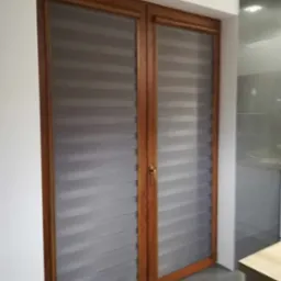 Dwu-skrzydłowe okno z zamontowanymi szarymi roletami w systemie dzień i noc, widoczne poziome pasy zaciemniające i przepuszczające światło, brązowa rama okienna.