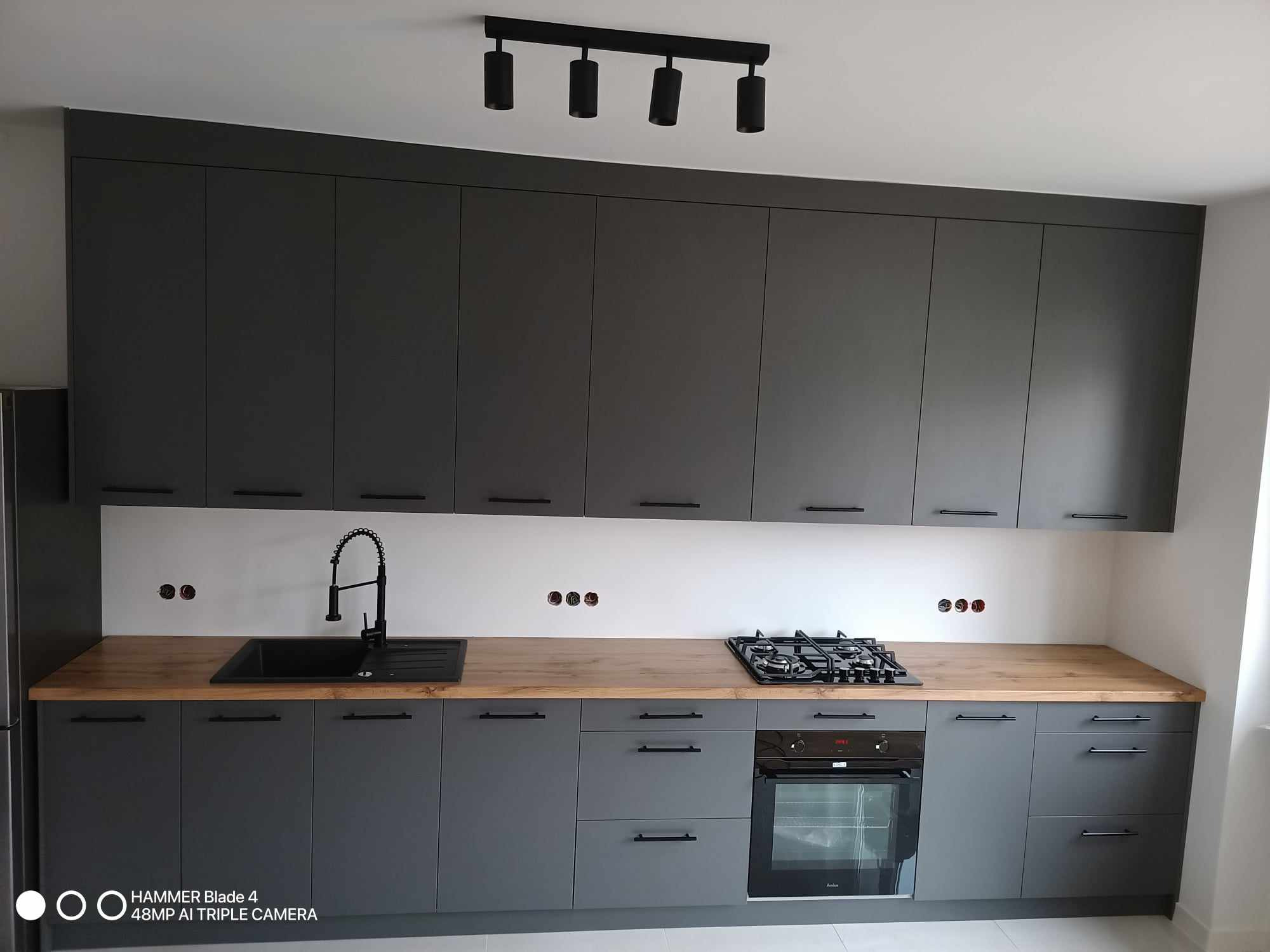 Nowoczesna kuchnia w odcieniach szarości z drewnianym blatem, czarną armaturą i oświetleniem punktowym, widoczne otwory na gniazdka elektryczne nad blatem.