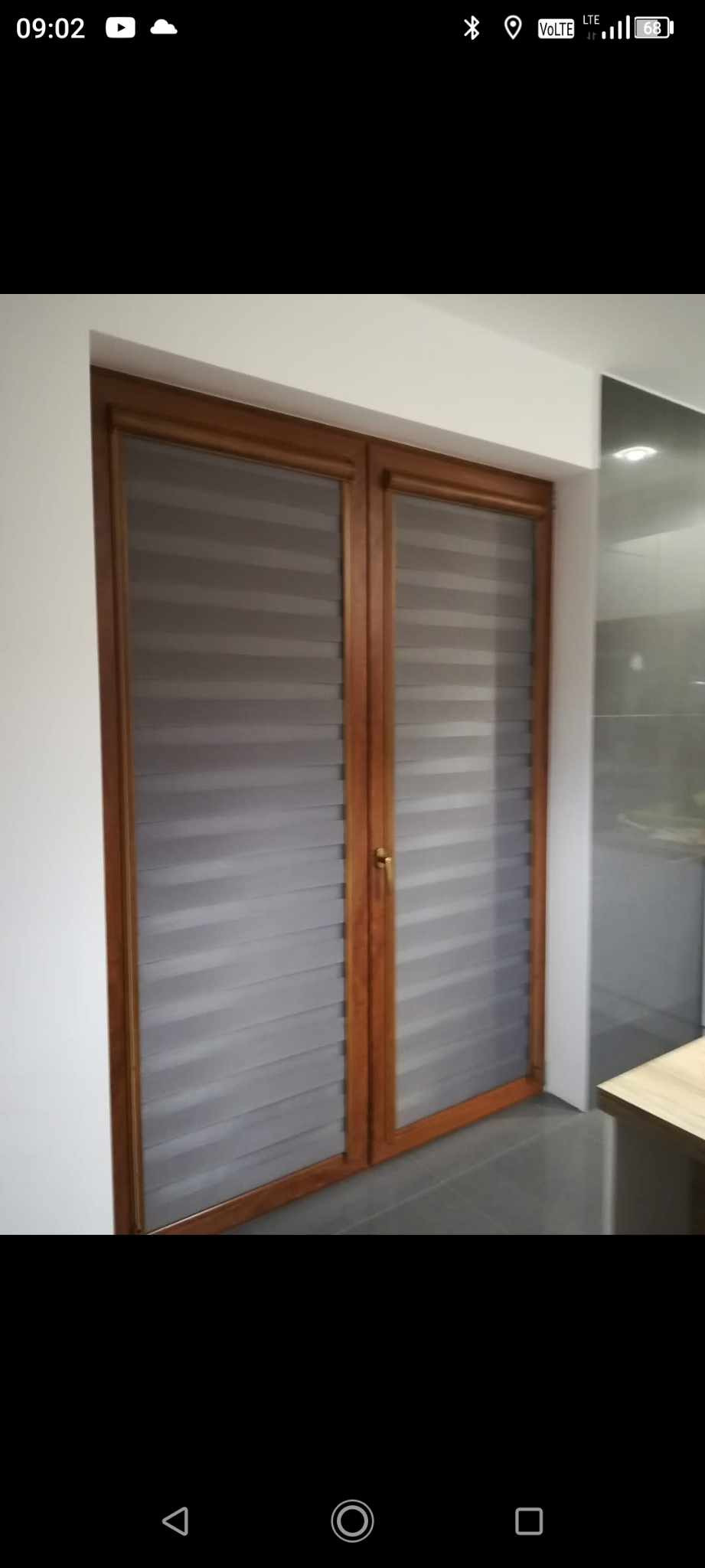 Dwu-skrzydłowe okno z zamontowanymi szarymi roletami w systemie dzień i noc, widoczne poziome pasy zaciemniające i przepuszczające światło, brązowa rama okienna.