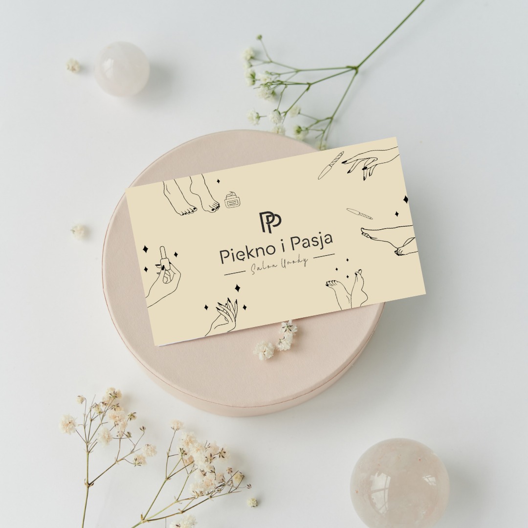 Kremowa wizytówka salonu urody z minimalistycznymi rysunkami dłoni i stóp, umieszczona na pudrowym talerzyku, otoczona kompozycją z gipsówki i dwóch kulistych kamieni.