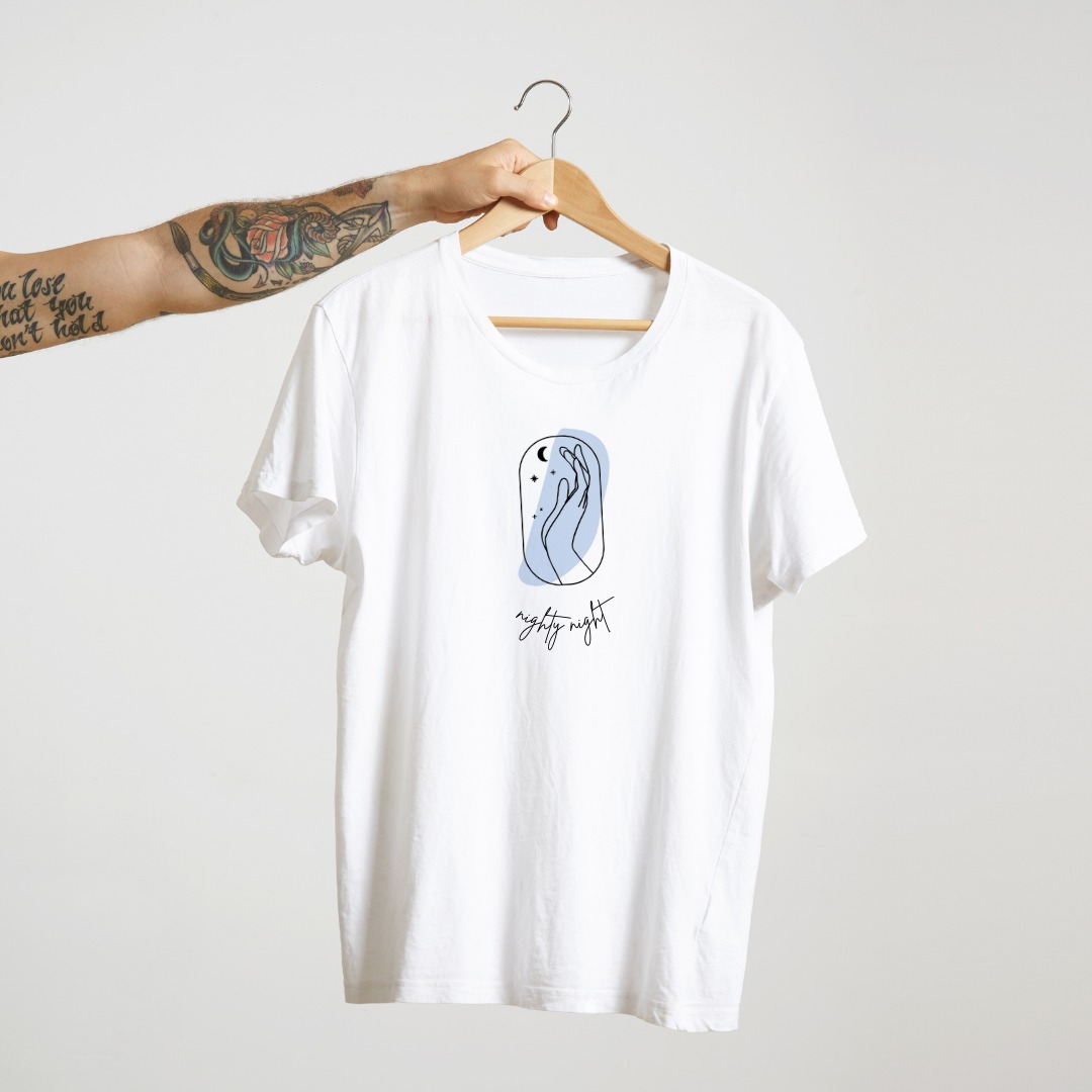 Biała koszulka z minimalistycznym nadrukiem przedstawiającym dłoń, księżyc i gwiazdy w delikatnej, błękitnej ramce, zawieszona na drewnianym wieszaku trzymanym przez wytatuowaną rękę.