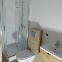 Nowoczesna łazienka z kabiną prysznicową, podwieszaną toaletą z zabudową w kolorze drewna i umywalką z szafką, wykończona białymi płytkami i panelami podłogowymi imitującymi drewno.