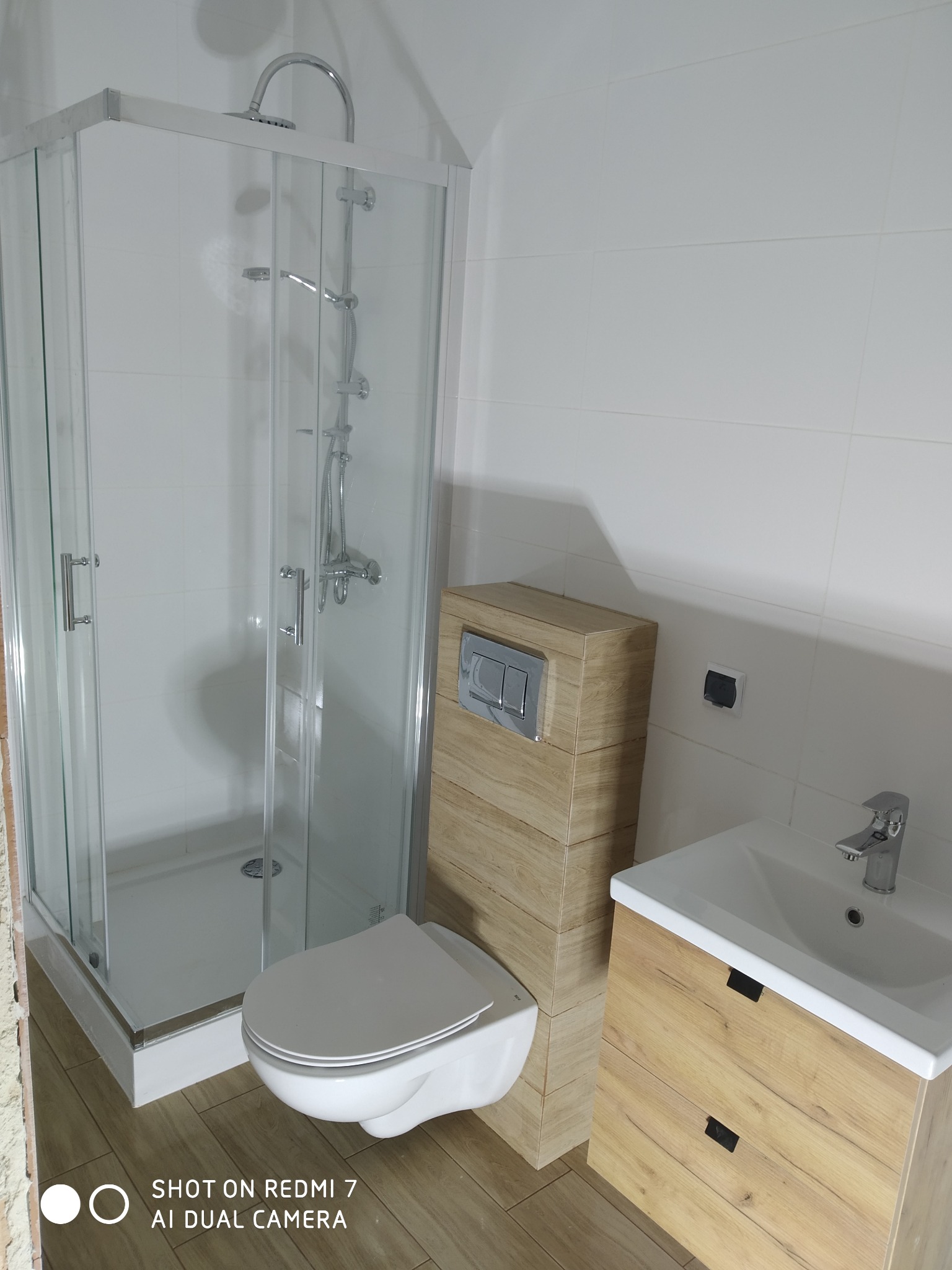 Nowoczesna łazienka z kabiną prysznicową, podwieszaną toaletą z zabudową w kolorze drewna i umywalką z szafką, wykończona białymi płytkami i panelami podłogowymi imitującymi drewno.