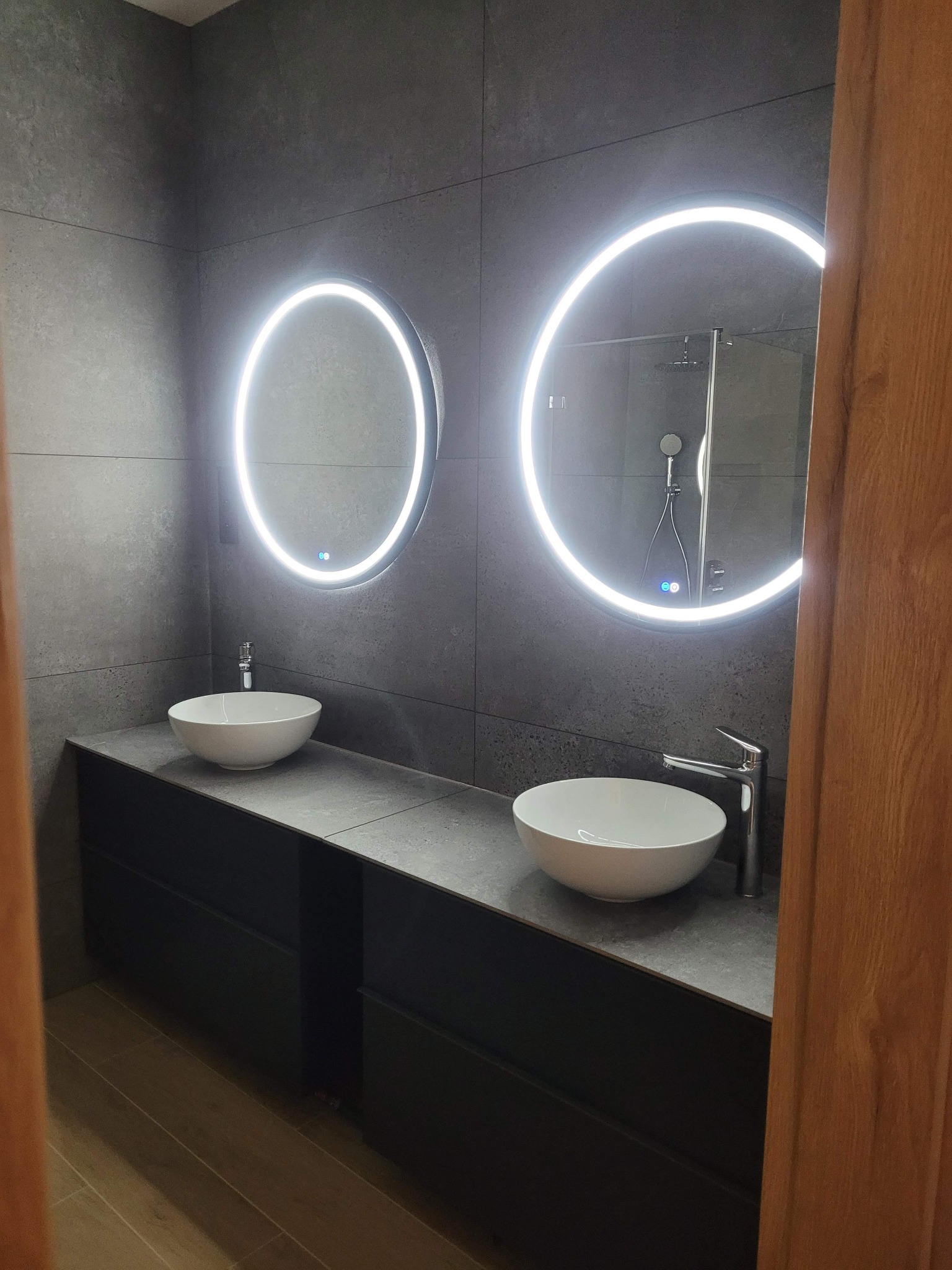 Nowoczesna łazienka z szarą płytką, dwoma okrągłymi lustrami z podświetleniem LED, dwiema białymi umywalkami nablatowymi i czarną szafką pod umywalki. W tle widoczny prysznic.