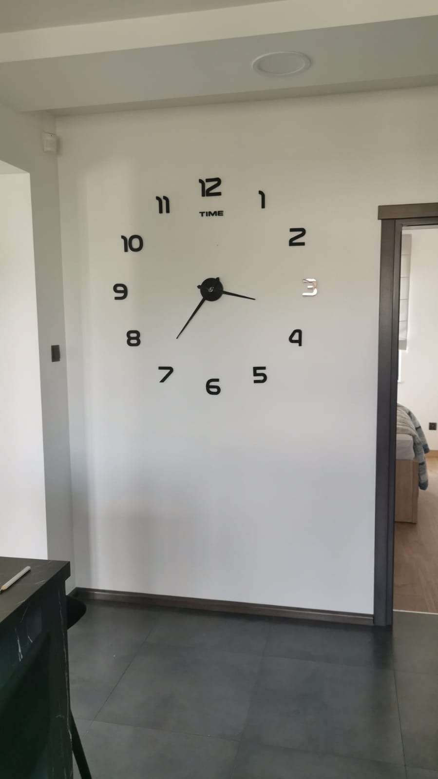 Biała ściana z dużym, czarnym zegarem naklejonym, wskazującym godzinę 7:40. Widoczny fragment podłogi z szarych płytek i fragment blatu z ołówkiem.