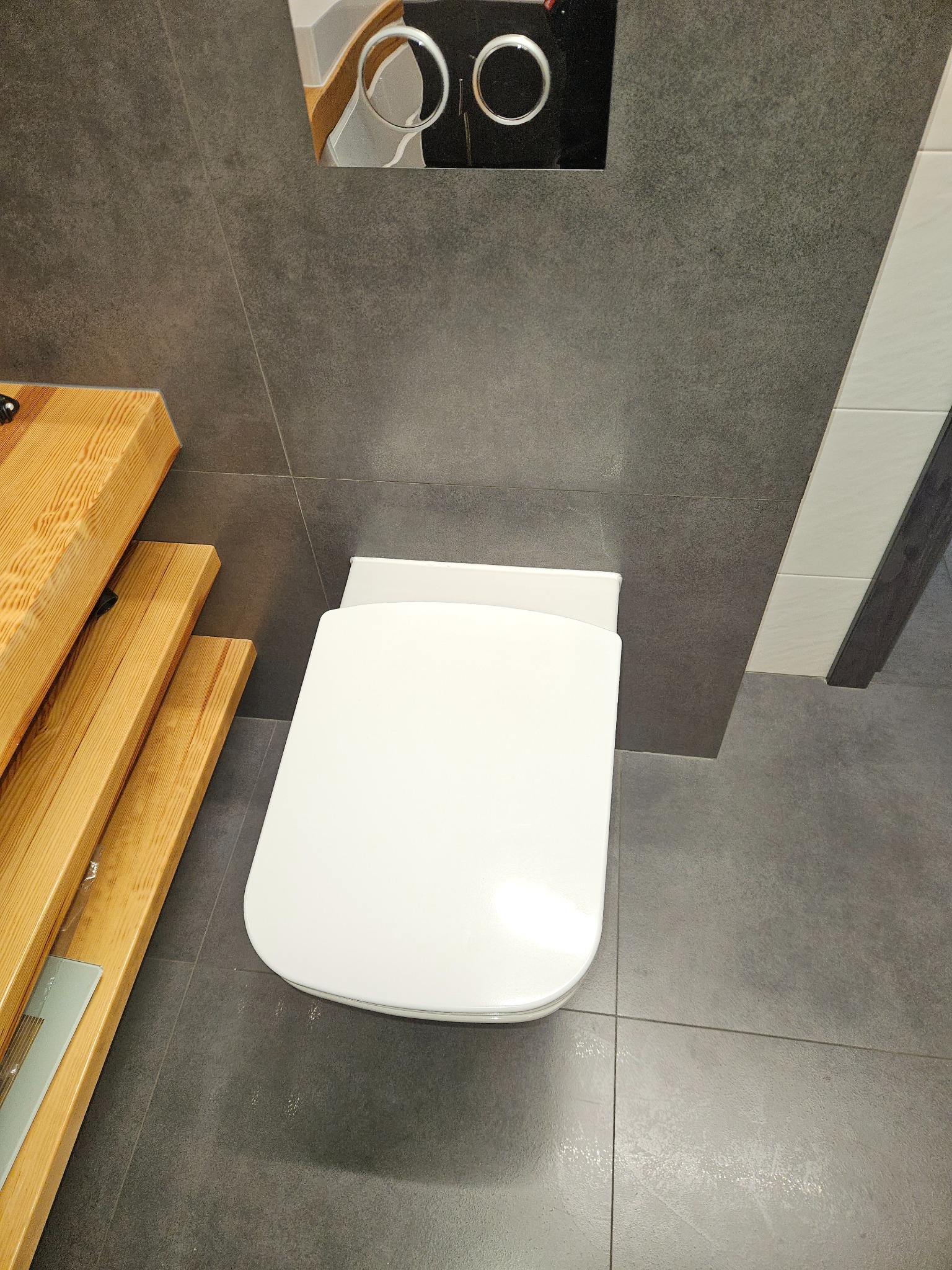 Nowoczesna łazienka z szarą, matową glazurą, drewnianymi akcentami i podwieszaną toaletą. Ujęcie z góry podkreśla minimalistyczny design i dbałość o detale.