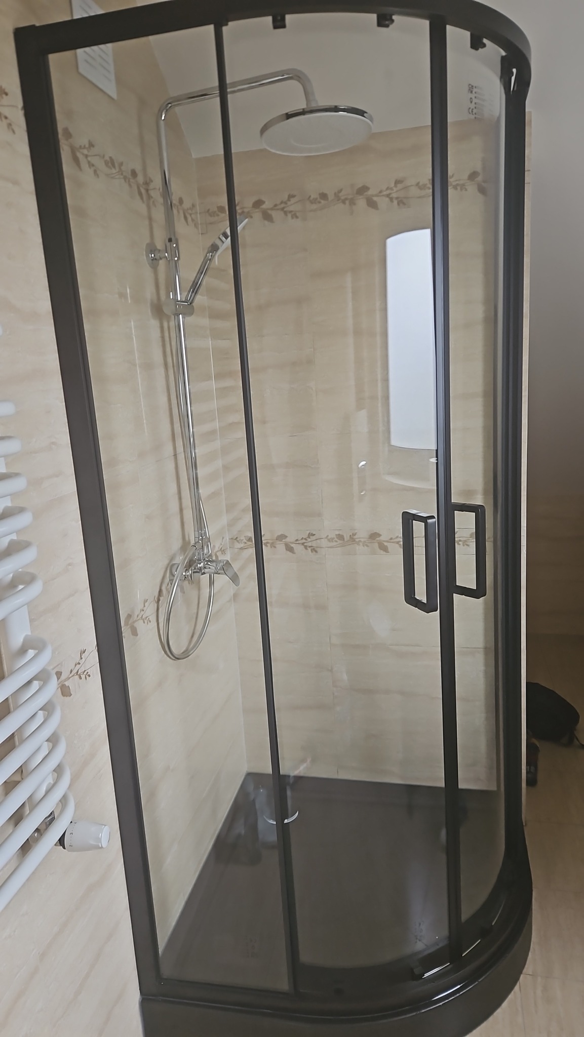 Nowoczesna kabina prysznicowa z czarną ramą i deszczownicą, zamontowana w łazience z beżowymi płytkami i grzejnikiem.